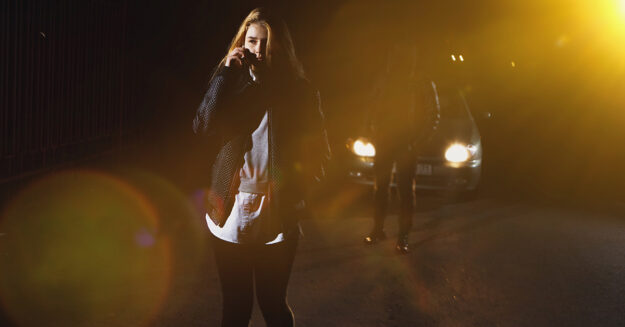 Is Uber Safe To Ride Alone female standing in the dark with light illuminating her from behind while she's on the phone a car is waiting with mysterious man behind her